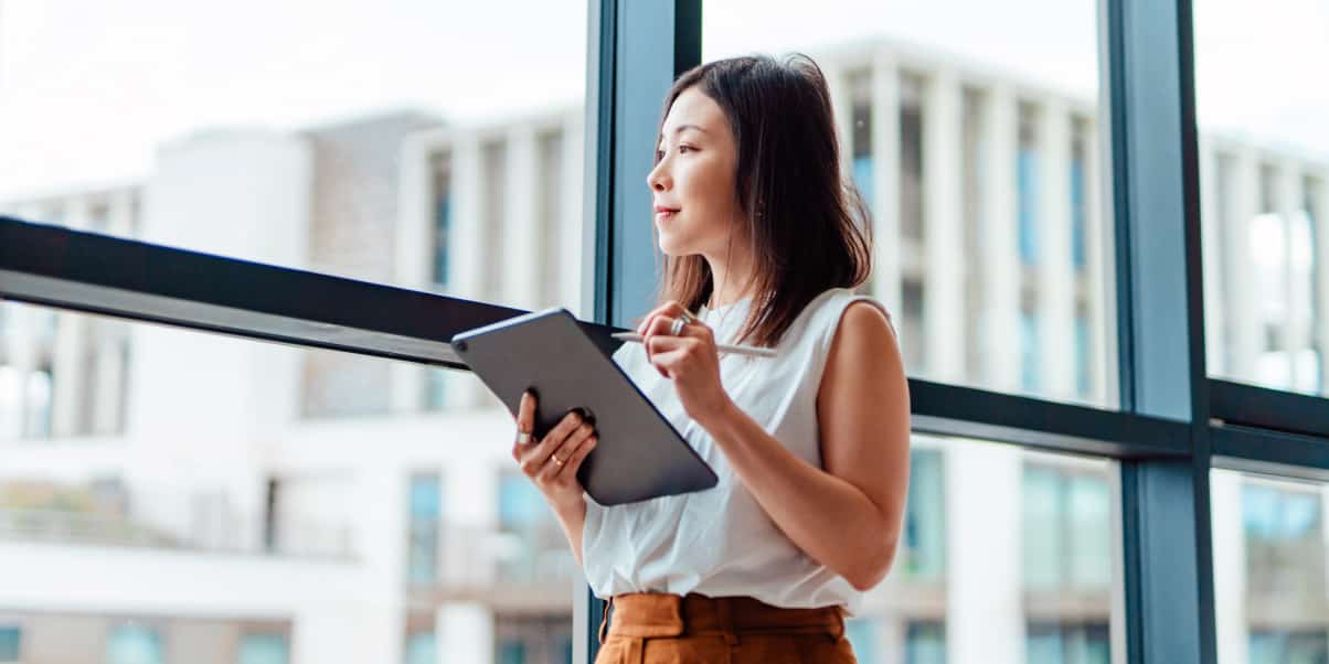 A woman looks out a large window while holding a tablet.